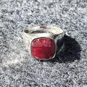May include: A silver ring with a square, deep red gemstone. The ring has a thick band and a textured appearance. The gemstone is set flush with the ring's surface. The image includes the text "JewelsArt".