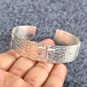 Peut inclure: Un bracelet manchette argenté avec une texture martelée et un motif en forme de croix. Le bracelet a un design ouvert et est présenté sur une main. Le bracelet est un bijou.
