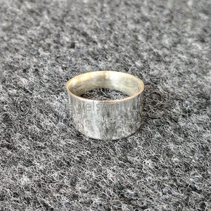 May include: A silver ring with a textured finish. The ring is a wide band design, with a simple, minimalist aesthetic. The ring is likely made of metal and is suitable for everyday wear.