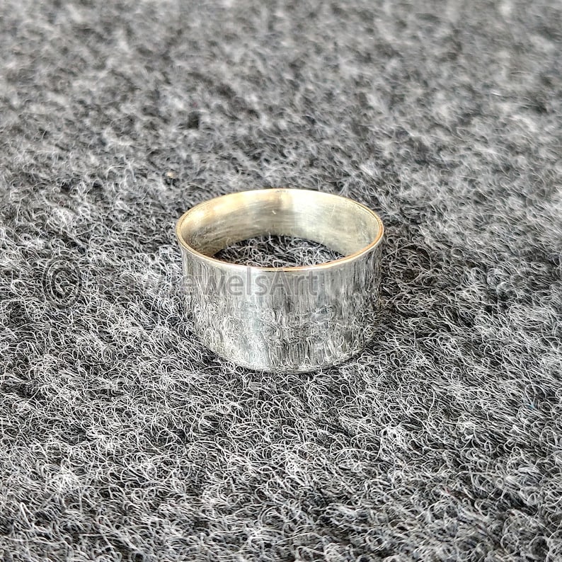 May include: A silver ring with a textured finish. The ring is a wide band design, with a simple, minimalist aesthetic. The ring is likely made of metal and is suitable for everyday wear.