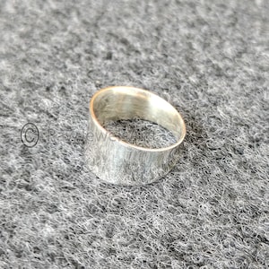 May include: A wide, silver ring with a textured finish. The ring has a simple, modern design and appears to be made of metal. The ring is displayed on a grey, textured surface.