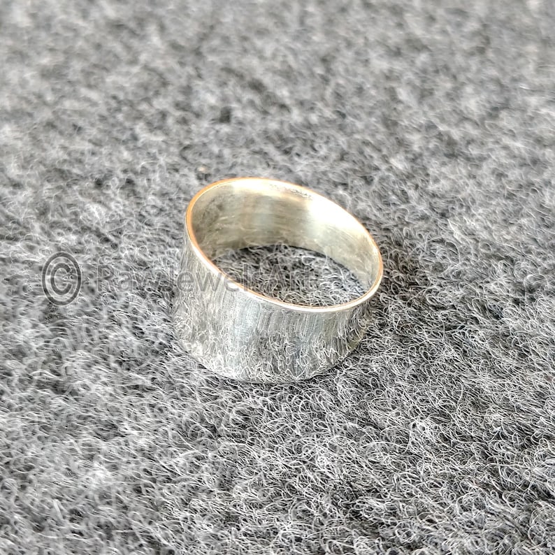 May include: A wide, silver ring with a textured finish. The ring has a simple, modern design and appears to be made of metal. The ring is displayed on a grey, textured surface.