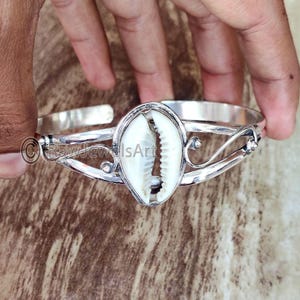 May include: Silver bracelet with a white cowrie shell charm. The bracelet has a delicate, ornate design.