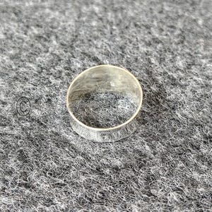 May include: A silver ring with a textured band. The ring is circular and rests on a grey, textured surface. The ring's band features a detailed, etched pattern around its circumference.