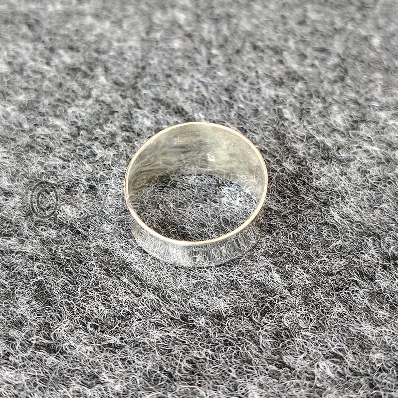 May include: A silver ring with a textured band. The ring is circular and rests on a grey, textured surface. The ring's band features a detailed, etched pattern around its circumference.