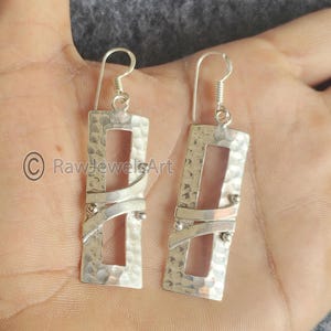 May include: A pair of silver rectangular earrings with a hammered texture and a cut-out design. The earrings feature a curved silver accent and small silver beads. The ear wires are a simple hook style.