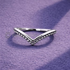Peut inclure: Une bague argentée en forme de V. La bague est ornée de petites perles rondes le long des bords, créant un aspect texturé. La bague est posée sur un fond violet.