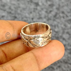 Peut inclure: Une bague en argent avec un motif animalier détaillé. La bague est large et présente une surface texturée avec des figures d'animaux sculptées. La bague est tenue par les doigts d'une personne sur un fond flou.
