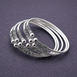 May include: A set of four silver-toned metal bangles featuring an open-ended design. Each bracelet is finished with a polished spherical bead at both ends, complemented by a textured, dark-oxidized section with intricate repeating patterns.
