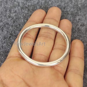 May include: A silver-colored, circular bangle bracelet. The smooth, polished metal reflects light, creating a bright appearance. The bangle is held in a hand, showcasing its simple, elegant design. The jewelry piece is a classic accessory.