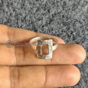 May include: A silver ring with a hammered square design. The ring has a rectangular cutout in the centre of the square. The ring is shown on a person's hand.