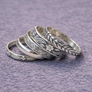 May include: A collection of silver rings with intricate floral and leaf patterns. The rings are stacked, showcasing the detailed designs. The rings are on a purple surface.