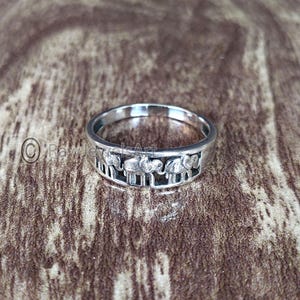Puede incluir: Un anillo de plata con un diseño de elefantes repetido. El anillo tiene una banda con formas rectangulares recortadas y los elefantes están en fila. El anillo está sobre una superficie marrón.