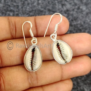 May include: Silver-tone drop earrings with cream-coloured cowrie shells in a teardrop shape. The shells have a natural texture and are set in a silver metal frame. The earrings have a simple hook design.