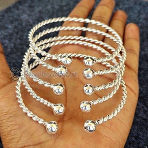 Könnte beinhalten: Fünf silberne Armreifen mit einem gedrehten Seil-Design und runden Kugelakzenten. Die Armreifen sind übereinander gestapelt.