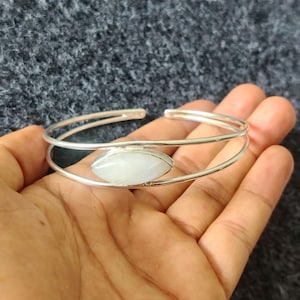 Könnte beinhalten: Ein silbernes Armreif-Armband mit einem ovalen, weißen Edelstein in der Mitte. Das Armband hat ein Doppelband-Design, wobei der Edelstein zwischen den Bändern eingesetzt ist. Das Armband ist hinten offen.