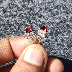 May include: A pair of silver stud earrings with vibrant red gemstones. The earrings are held up against a gray background. The gemstones are rectangular and set in a silver metal setting.