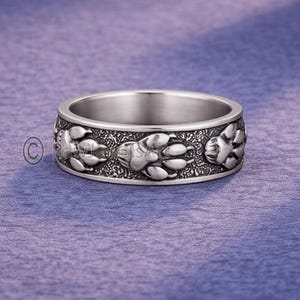 May include: A silver ring with a repeating paw print pattern. The band has a textured surface with raised paw prints. The ring is set against a purple background.