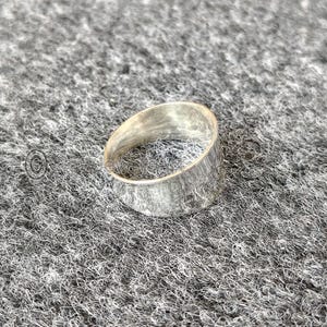 May include: A silver ring with a textured surface. The ring has a wide band and a simple, minimalist design. The ring is made of a shiny, metallic material.