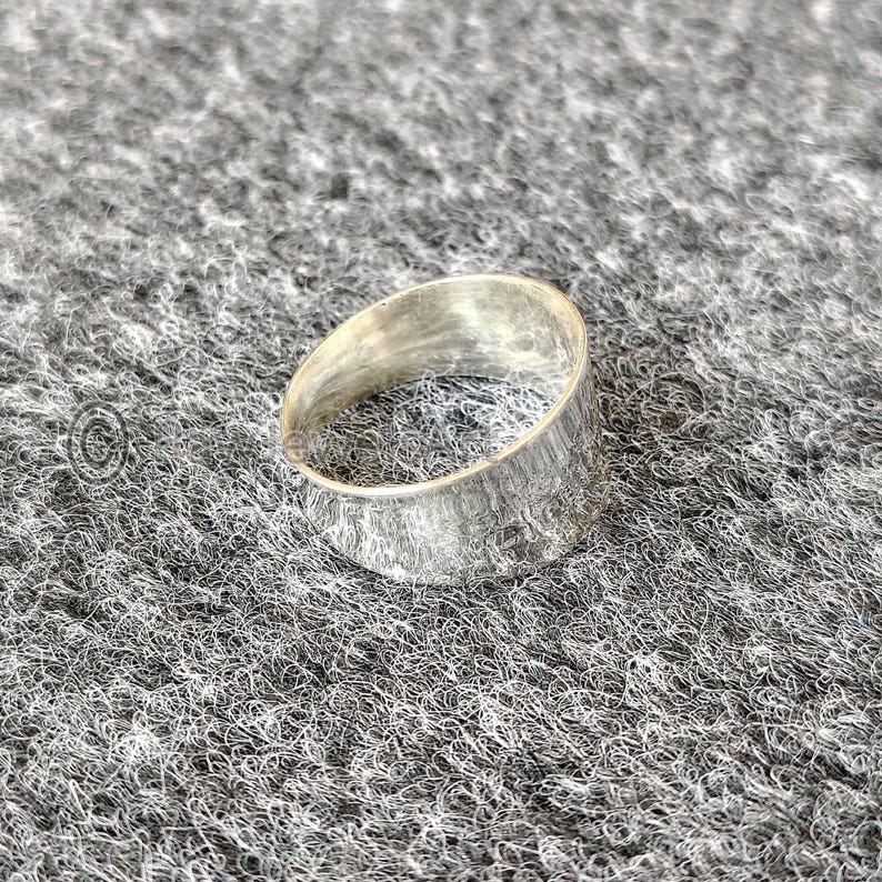 May include: A silver ring with a textured surface. The ring has a wide band and a simple, minimalist design. The ring is made of a shiny, metallic material.