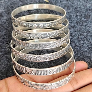 Könnte beinhalten: Ein Stapel silberner Armreifen mit aufwendigen floralen und geometrischen Mustern. Die Armreifen werden auf einer Hand präsentiert, wobei die detaillierten Designs und die reflektierende Qualität des Metalls hervorgehoben werden. Traditioneller Schmuck.