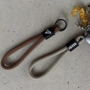 Könnte beinhalten: Zwei Schlüsselanhänger aus geflochtenem Seil, einer in Braun und einer in Beige. Jeder verfügt über einen schwarzen Metallverschluss mit weißem Aufdruck: ein Herz auf dem braunen Seil und der Schriftzug "kleani." auf dem beigen Seil. Ein goldener Harzanhänger ist befestigt.