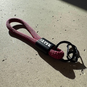 May include: A burgundy rope keychain with a black metal clasp and a black cylindrical piece with the word "keani." The keychain is on a light gray concrete surface.
