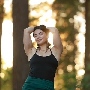 May include: A woman with her eyes closed, wearing a black tank top and a green skirt, stands in a forest with the sun shining through the trees.