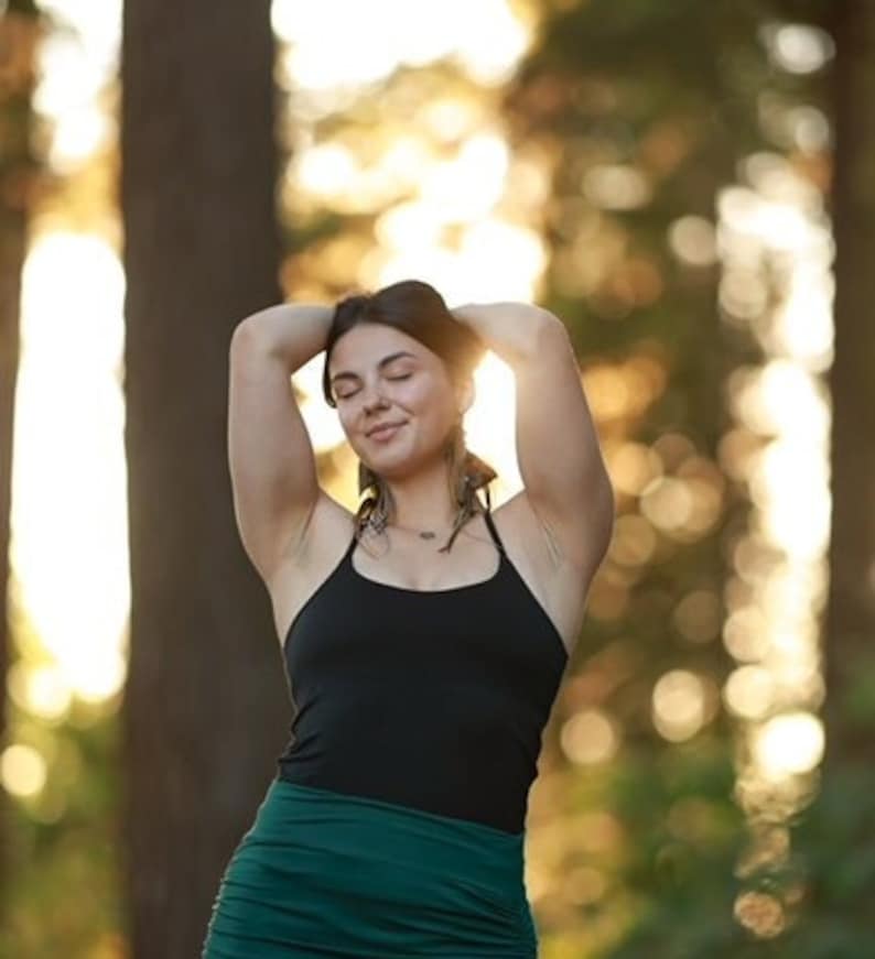 May include: A woman with her eyes closed, wearing a black tank top and a green skirt, stands in a forest with the sun shining through the trees.