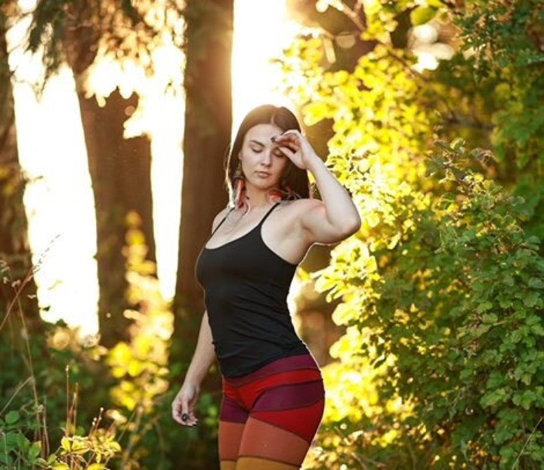 May include: A woman wearing a black tank top and colorful striped pants stands in a forest. The sun shines through the trees behind her.