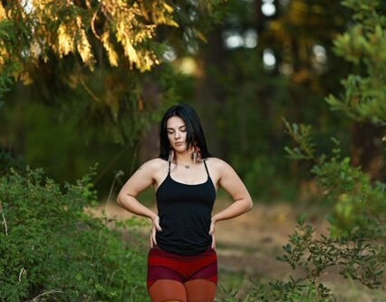May include: A woman with long black hair wearing a black tank top and red and orange patterned shorts stands in a forest setting.