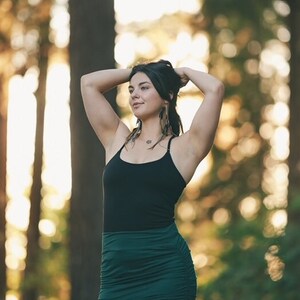 May include: A woman with long dark hair wearing a black tank top and a dark green skirt. She is standing in a forest with trees in the background.