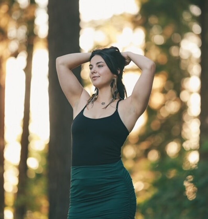 May include: A woman with long dark hair wearing a black tank top and a dark green skirt. She is standing in a forest with trees in the background.