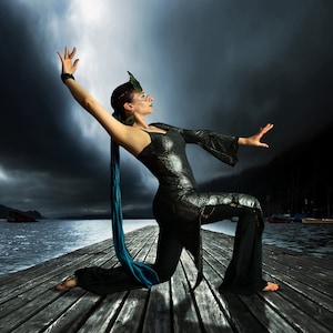 May include: A woman in a black and silver costume with a blue and green feather headdress poses on a wooden dock. She is reaching out with her right hand and her left hand is on her hip. The background is a dark stormy sky and a body of water.