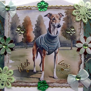 Op de afbeelding: Een rechthoekige wenskaart met een bruin-witte hond in een grijze gebreide trui, staand in een park. De kaart is versierd met groene kanten bloemen, lichtblauwe lintstrikjes, groene strass-steentjes en de gouden tekst With Love.