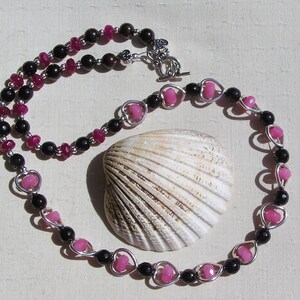 May include: A handmade necklace with black and pink beads, and heart-shaped accents. The necklace is displayed with a seashell on a light-coloured surface. The necklace has a silver clasp.