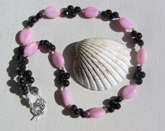 Puede incluir: Un collar de cuentas rosa y negro con un cierre plateado. El collar está hecho con cuentas rosadas ovaladas y pequeñas cuentas negras.