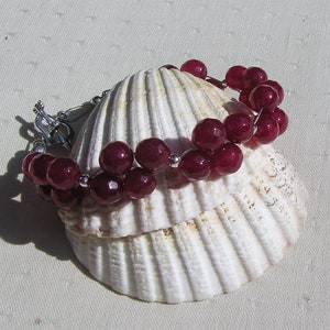 May include: A red gemstone bracelet with silver clasps and beads. The bracelet is displayed on a white seashell.