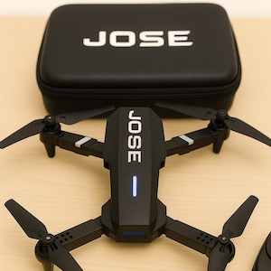 Puede incluir: Dron negro con cuatro hélices y el nombre "JOSE" impreso en la parte superior. El dron está sobre una superficie de madera junto a un estuche negro con "JOSE" impreso. El dron tiene una luz azul.