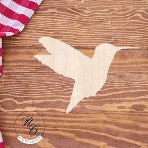 May include: A wooden hummingbird cutout in a flying pose, set against a brown wood grain background. A red and white checkered cloth is in the upper left corner. The logo for RC Canvas Cuts is in the lower center.