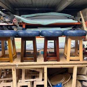 May include: Four wooden stools with blue leather seats. The stools have different wood finishes: light, medium, and dark brown. The stools are displayed on a wooden workbench in a workshop setting.