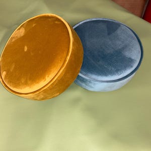 May include: Two round, velvet boxes. One is a golden-yellow colour, and the other is a muted teal blue. The boxes have a soft, plush texture and a simple, elegant design. They are likely used for storage or decoration.