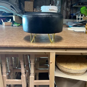 Puede incluir: Un otomán redondo negro con patas de horquilla doradas. El otomán está hecho de un material liso y oscuro y se eleva sobre cuatro patas anguladas. La pieza está sobre una mesa de madera en un taller.