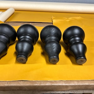 Puede incluir: Cuatro patas de muebles de madera negra con un diseño torneado. Cada pata presenta una parte superior redondeada, una sección central bulbosa y una base cónica. Las patas están dispuestas sobre una superficie amarilla, lo que sugiere un contexto de fabricación de muebles.