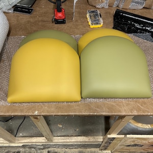 Puede incluir: Cuatro cojines de asiento semicirculares en tonos amarillo y verde están dispuestos sobre una superficie de madera. Los cojines son probablemente para sillas o taburetes. En el fondo se ven herramientas y materiales de embalaje.
