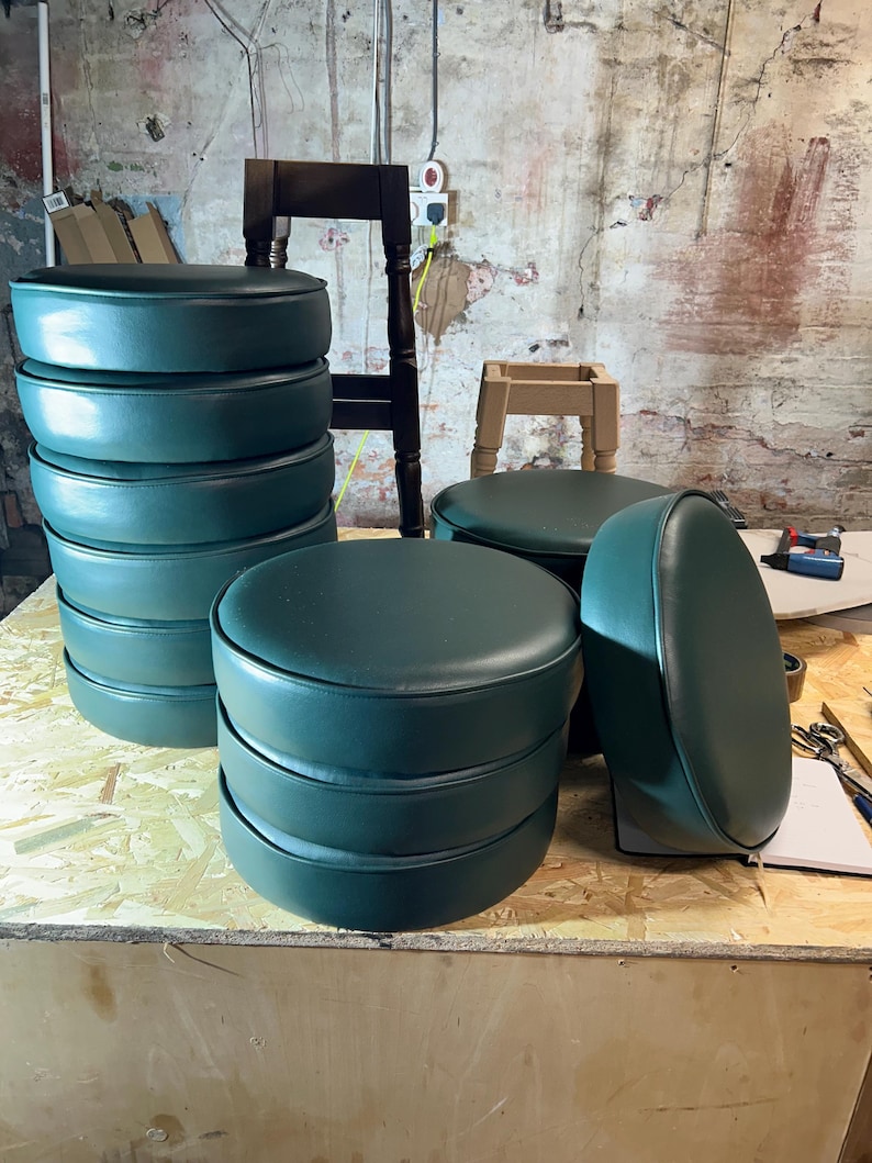 Puede incluir: Una pila de taburetes de bar redondos de color verde oscuro con base de madera. Los taburetes est&aacute;n apilados sobre una superficie de madera.