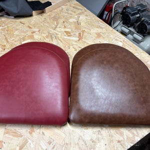 Puede incluir: Dos cojines de silla, uno rojo y otro marrón, se muestran sobre una superficie de madera. Los cojines están hechos de una tela texturizada y tienen una forma redondeada.