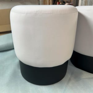 May include: Two white upholstered ottomans with a black base. The ottomans have a rounded, cylindrical shape. The background includes a cardboard box and a light blue surface.