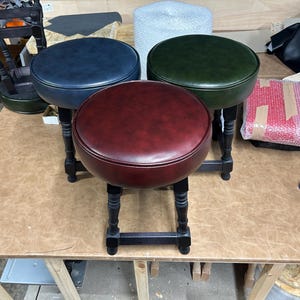 May include: Four round stools with dark wooden legs and padded seats. The seats are in shades of blue, green, and burgundy. The stools are on a light brown surface.