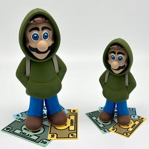 May include: Two figurines of a cartoon character wearing a green hoodie, blue pants, and brown shoes. The figurines stand on colorful, stylized money. The larger figure is in front of the smaller one. The figures have a mustache and blue eyes.